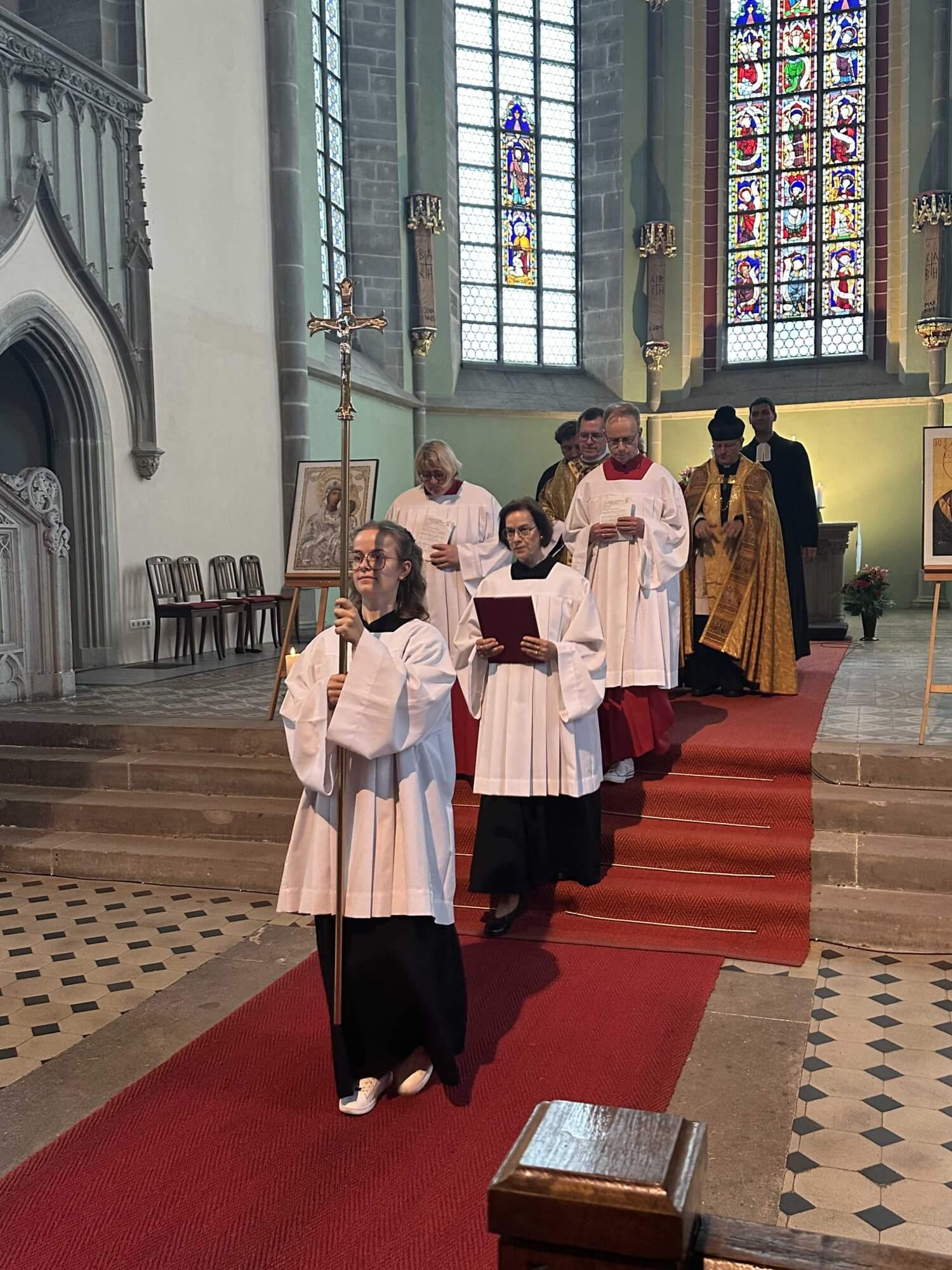 Eine Gruppe von Menschen zieht in Messgewändern aus dem Altarraum einer Kirche aus. Die vorderste Person trägt ein Vortragekreuz aus Gold. Hinter ihr läuft ein Mann mit einem goldenen Evangeliar. Im Hintergrund sind weitere Amtsträger zu erkennen