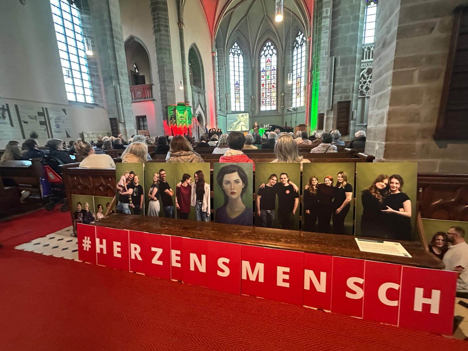 Man sieht eine vollbesetzte Kirche im Vordergrund stehen Bildtafeln auf einer Bank. Darunter ist #Herzensmensch zu lesen.