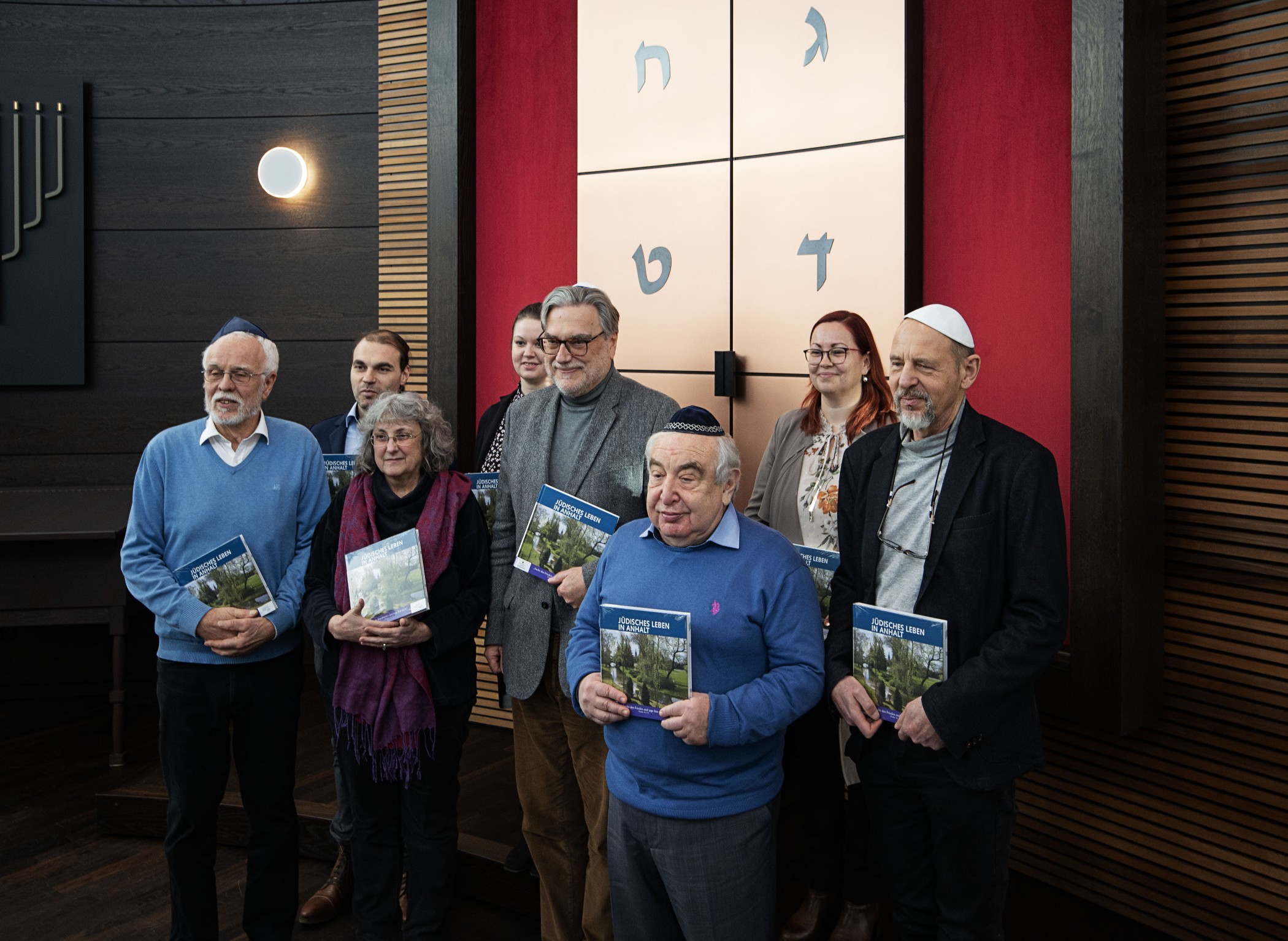 Auf dem Bild sind 8 Personen zu sehen. Drei Frauen und 5 Männer. Die Männer tragen eine Kippa. Alle stehen vor einer Wand mit hebräischen Buchstaben und halten ein Buch in der Hand. 