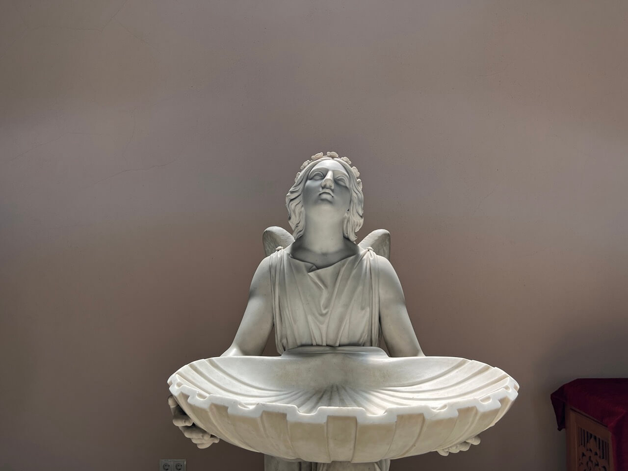 ein Taufengel aus weißem Marmor der eine große Jakobsmuschel als Taufbecken hält.