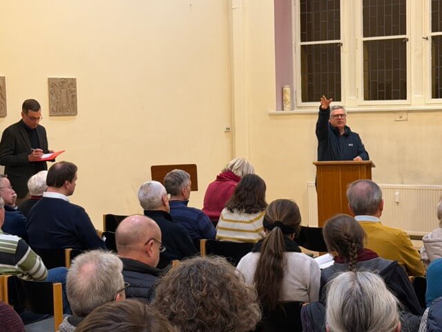 Auf dem Bild sind viele Menschen in einem Saal zu sehen. Sie sitzen in Richtung des Rednerpultes, das vorne links steht. An dem Pult steht ein Mann der mit der Hand in Richtung der Kamera deutet.