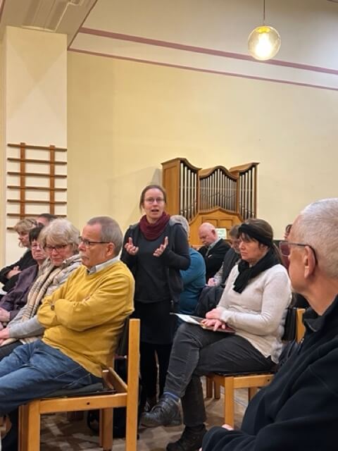 Im Mittelpunkt steht eine junge Frau, die gerade redet inmitten von sitzenden Menschen