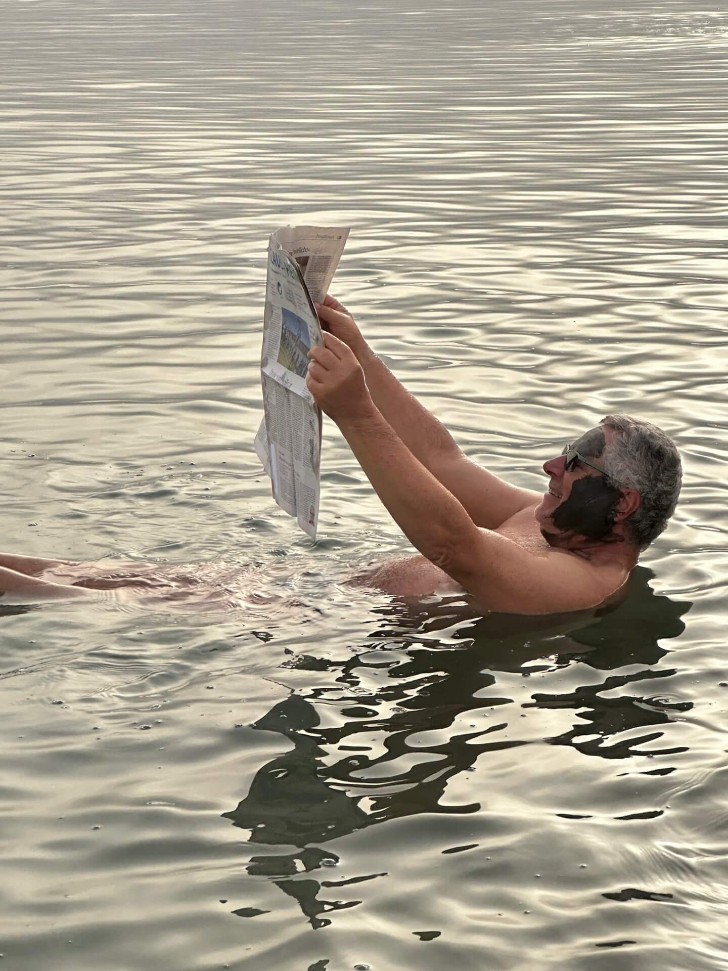 Ein Mann mit Sonnenbrille und einer aufgeschlagenen Zeitung schwimmt auf dem Rücken liegend auf dem toten Meer. 