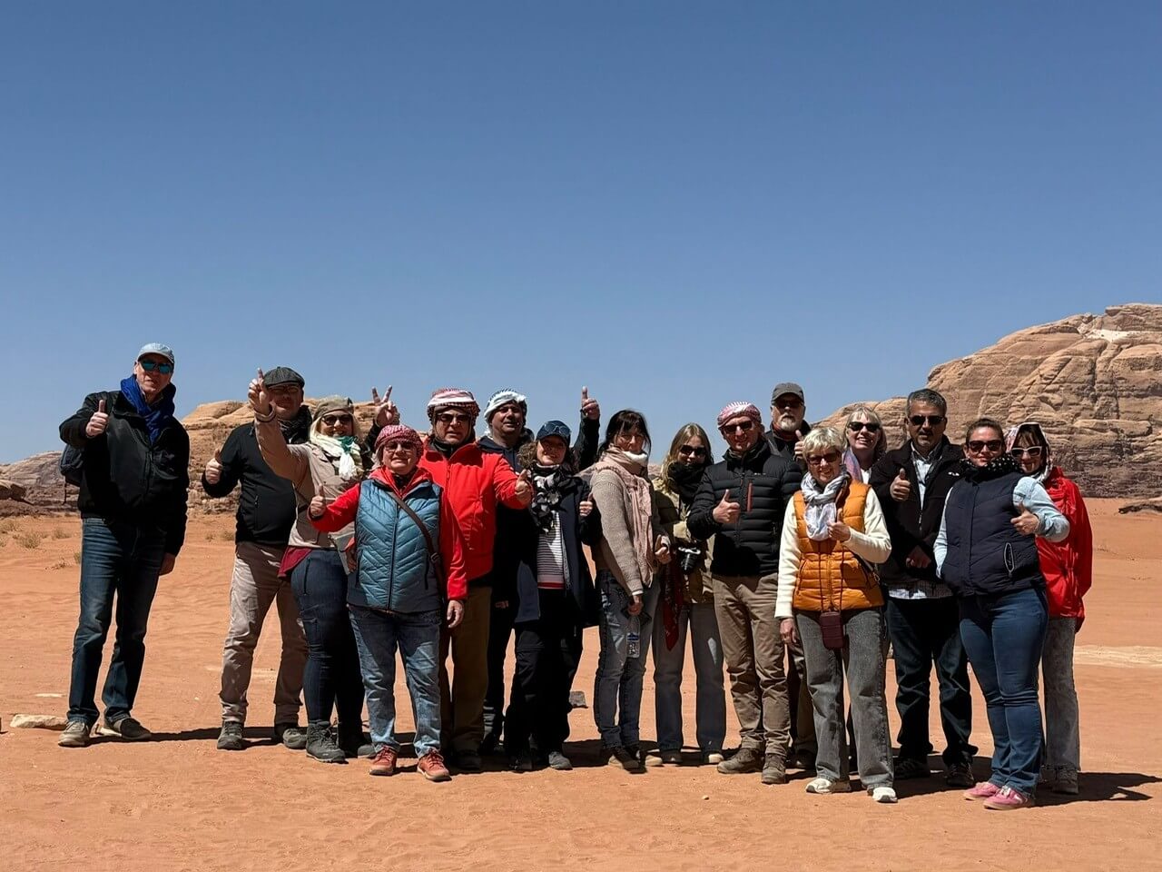 Eine Gruppe von Menschen steht auf dem Sand in der Jordanischen Wüste. Der Sand ist orange-rötlich, der Himmel darüber ganz blau ohne eine Wolke