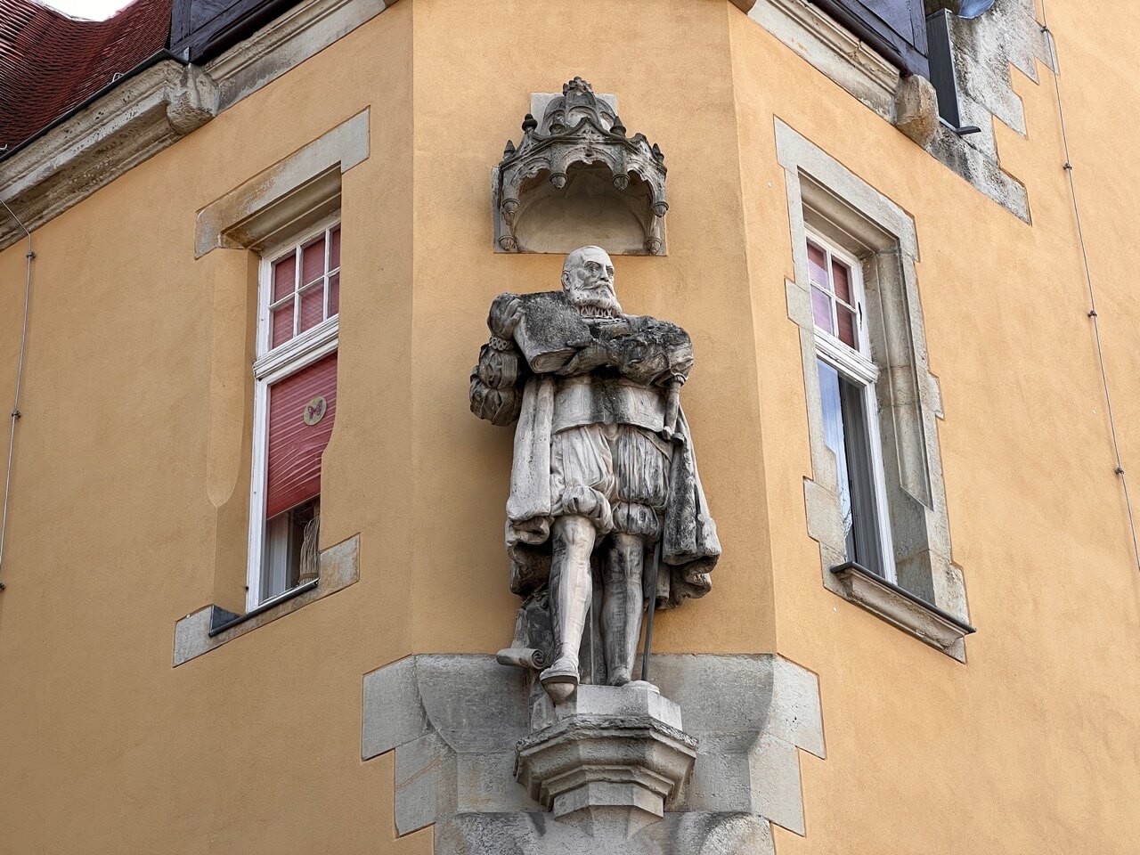 Auf dem Bild ist eine Statue des Fürsten Wolfgang zu sehen, die ein Buch in der Hand hält und in die Ecke eines gelben Gebäudes eingelassen ist.