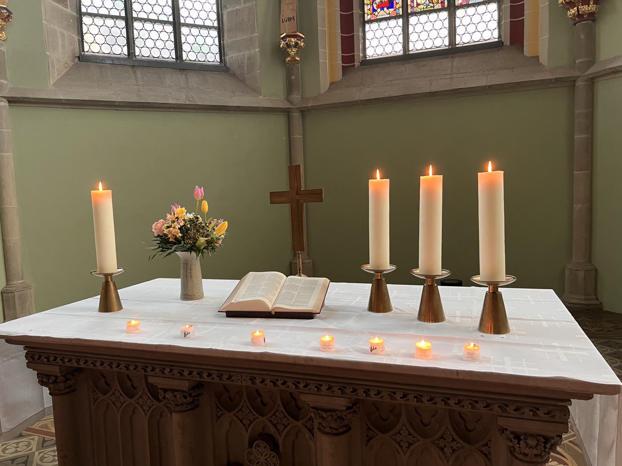 Bild von einem Altar, darauf stehen von links nach rechts: eine Kerze, ein Blumenstrauß, ein Kreuz und drei Kerzen. Davor stehen brennende Teelichter auf dem weißen Tischtuch