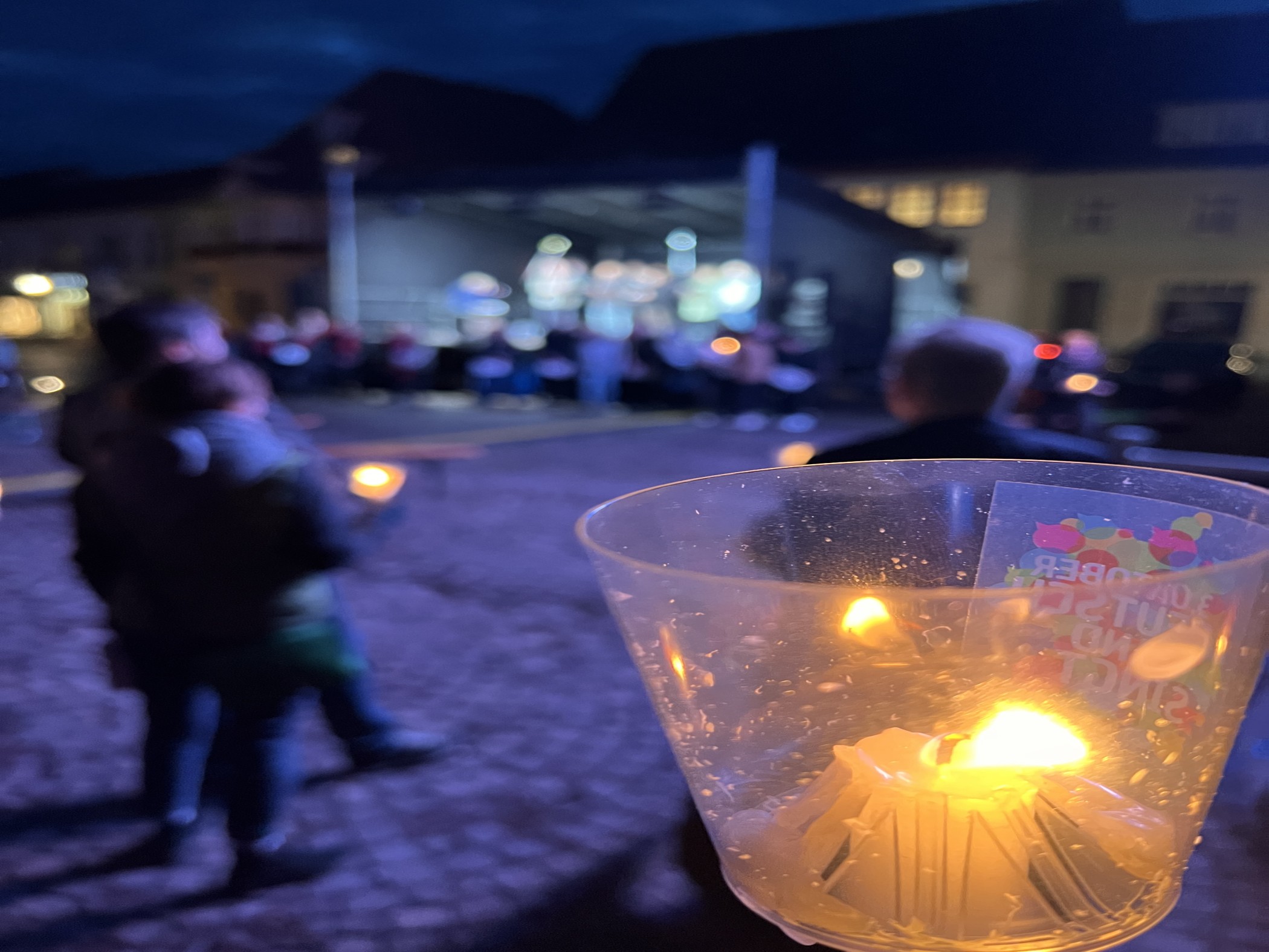 Im Vordergrund eine Kerze in einem Windlicht, dahinter Menschen die Kerzen halten auf dem Marktplatz im Dunkeln