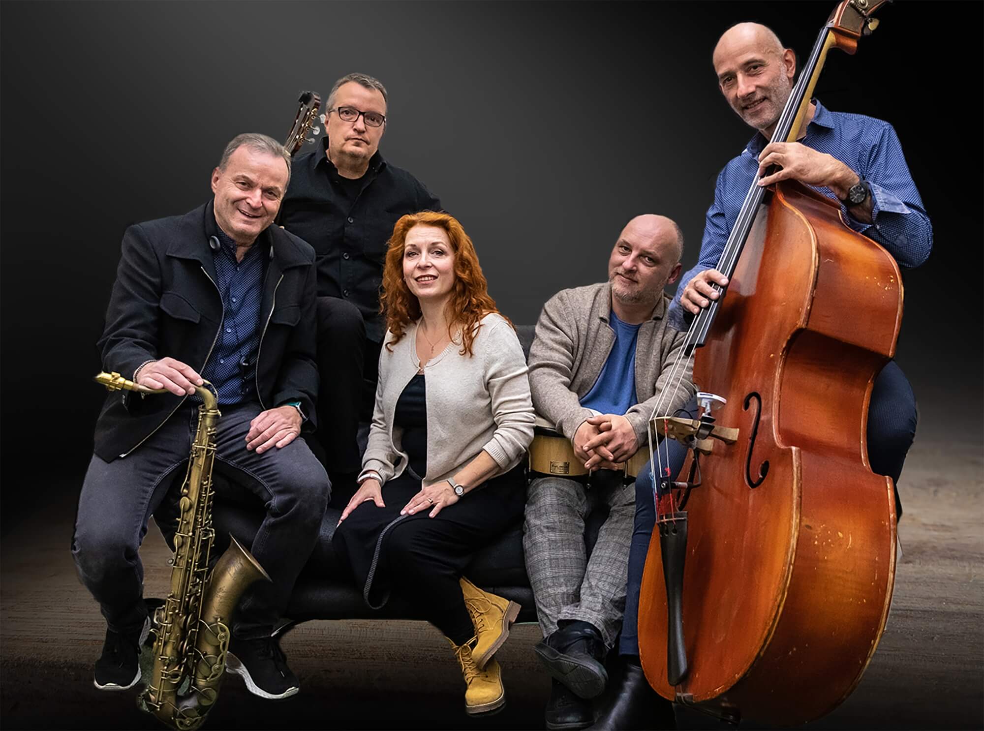 Bandfoto der Band Danca a Bossa. Zu sehen ist von links nach rechts: ein Mann in schwarzer Jacke der sich auf ein Saxofon stützt, im Hintergrund ein schwarz gekleideter Mann mit E-Gitarre in der Hand, im Vordergrund eine Frau mit grauem Oberteil, daneben ein Mann mit beiger Jacker, der zwei Trommeln auf dem Schoß hat und ganz rechts ein Mann mit blauem Hemd der hinter einem Kontrabass steht.