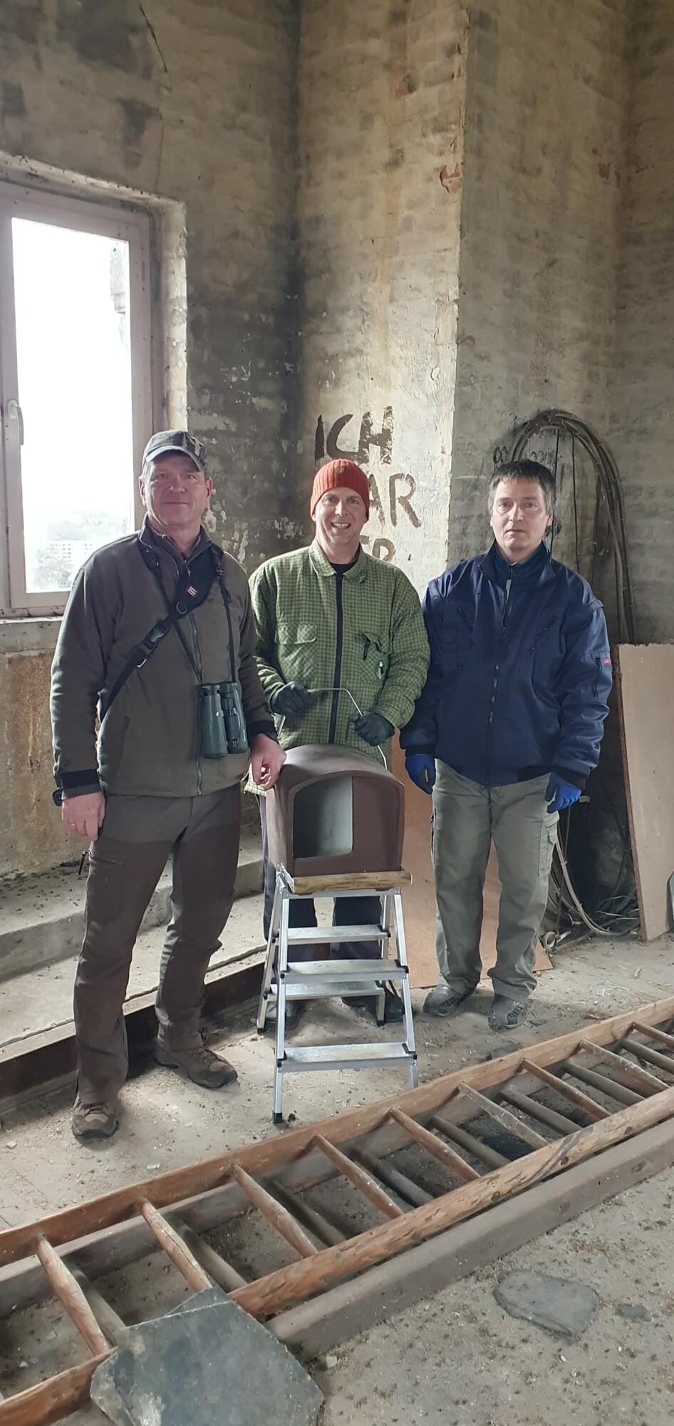 Auf dem Bild sind drei Männer hinter einem Turmfalkenkasten zu sehen. Sie stehen im Inneren des Turmes und im Hintergrund sieht man ein Fenster.