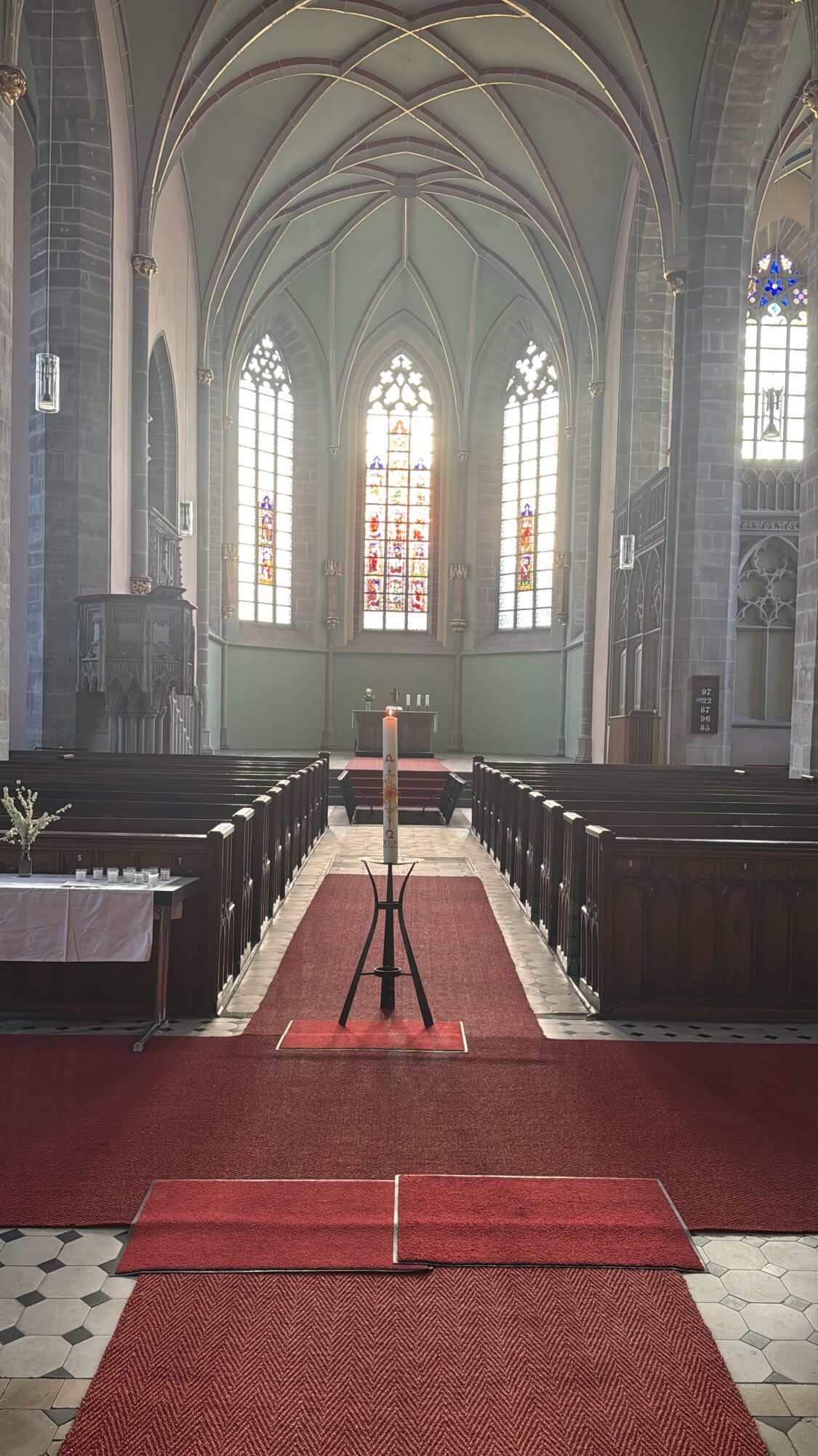 Eine brennende Osterkerze steht auf einem Ständer in einer leeren Kirche. Der Blick ist Richtung Altarraum und durch die Fenster dahinter scheint die Sonne herein. 