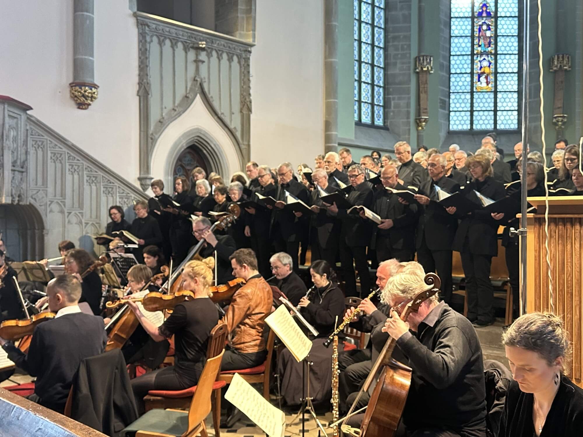 Blick von der Seite auf ein Orchester hinter dem ein großer Chor steht. Alle sind schwarz gekleidet und im Hintergrund ist zu erkennen, dass es in einer Kirche stattfindet.
