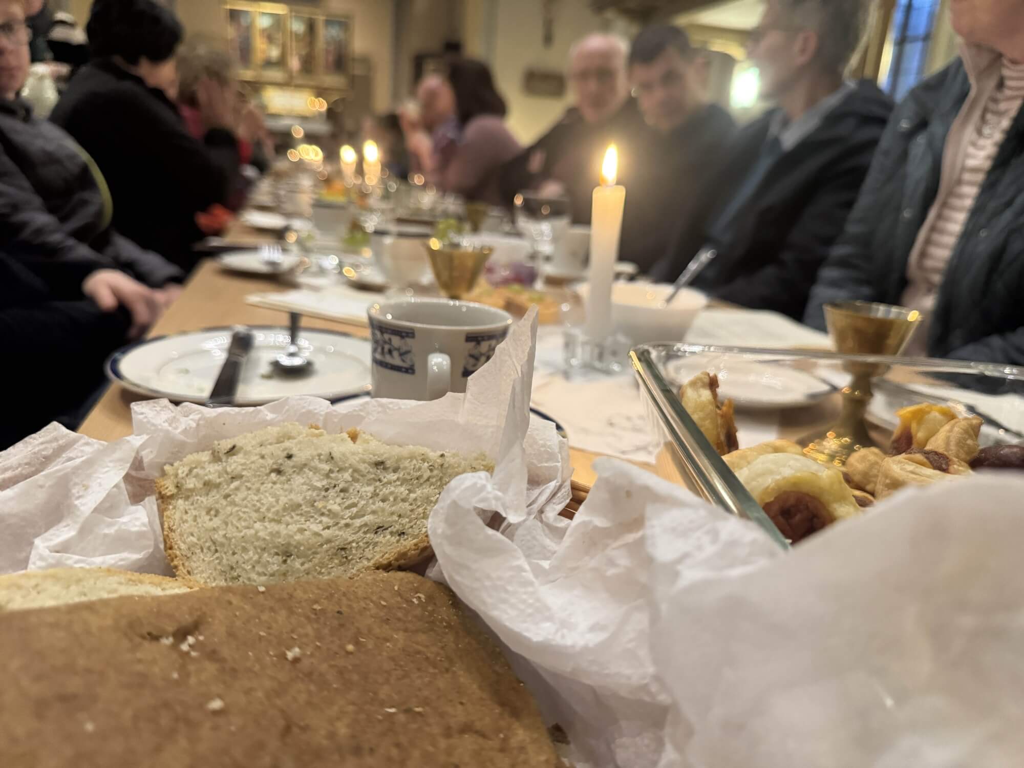 Blick auf einen Tisch an dem viele Menschen sitzen. Auf dem Tisch stehen Kerzen, Teller, Tassen und Brot. Im Hintergrund ist der Altar zu sehen.