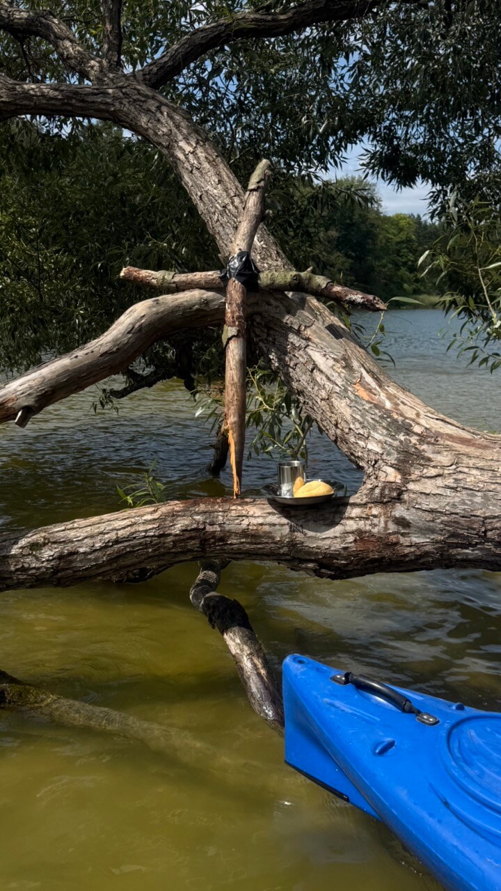 Ein Holzkreuz, das mit Klebeband zusammengehalten wird, steht auf einem liegenden Baum im Wasser. Davor stehen ein silberner Becher und ein Teller mit einem Brot.
