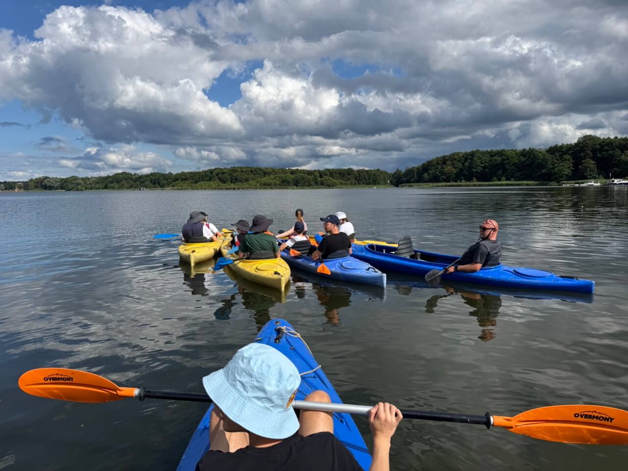 Menschen in gelben und blauen Kajaks liegen mitten auf einem See beieinander und beraten.