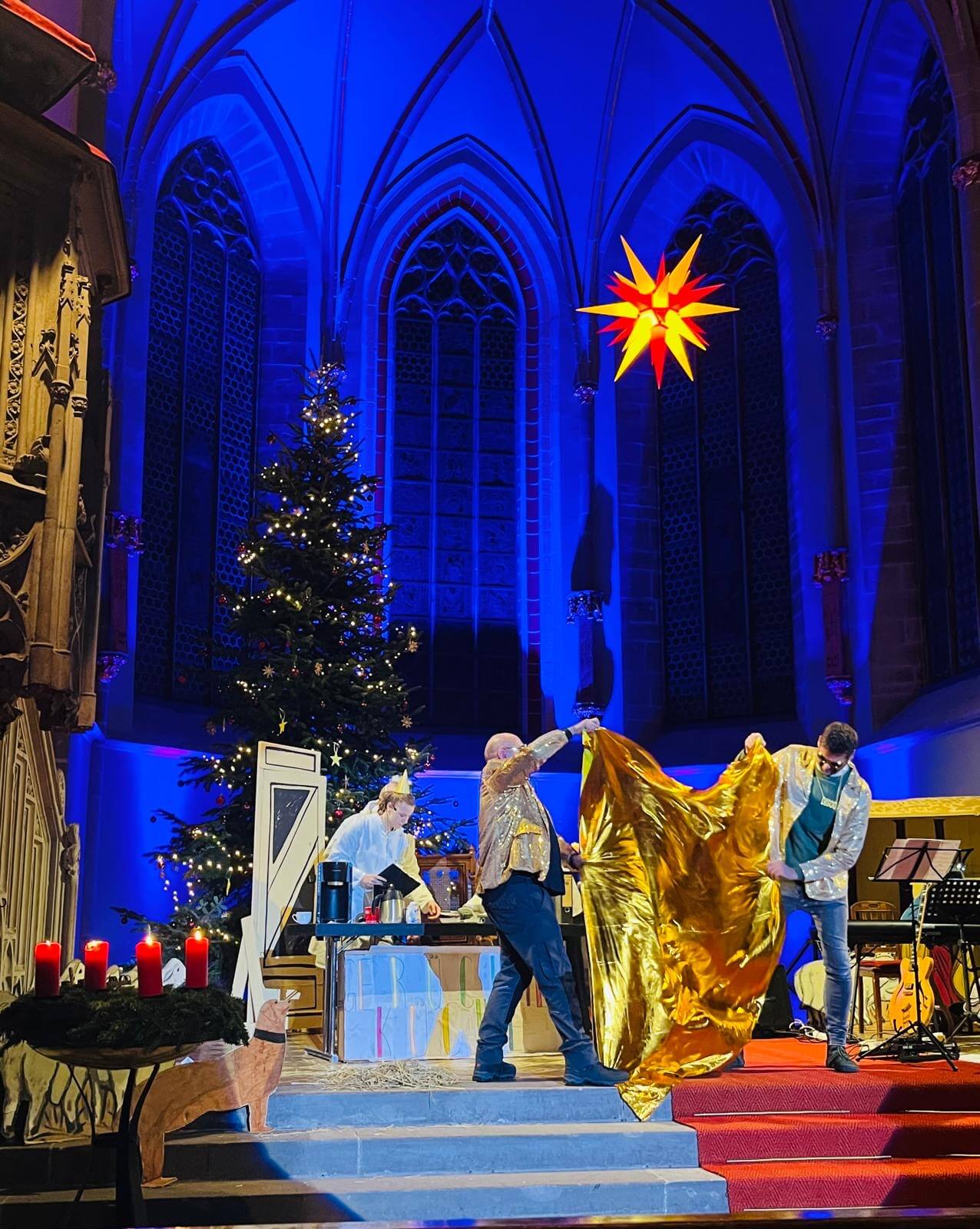 Auf dem Bild ist der Altarraum der Kirche zu sehen. Ein Weihnachtsbaum steht geschmückt im Hintergrund. Vorne stehen zwei Personen mit glitzernden Anzügen, die ein glitzerndes Tuch halten. 