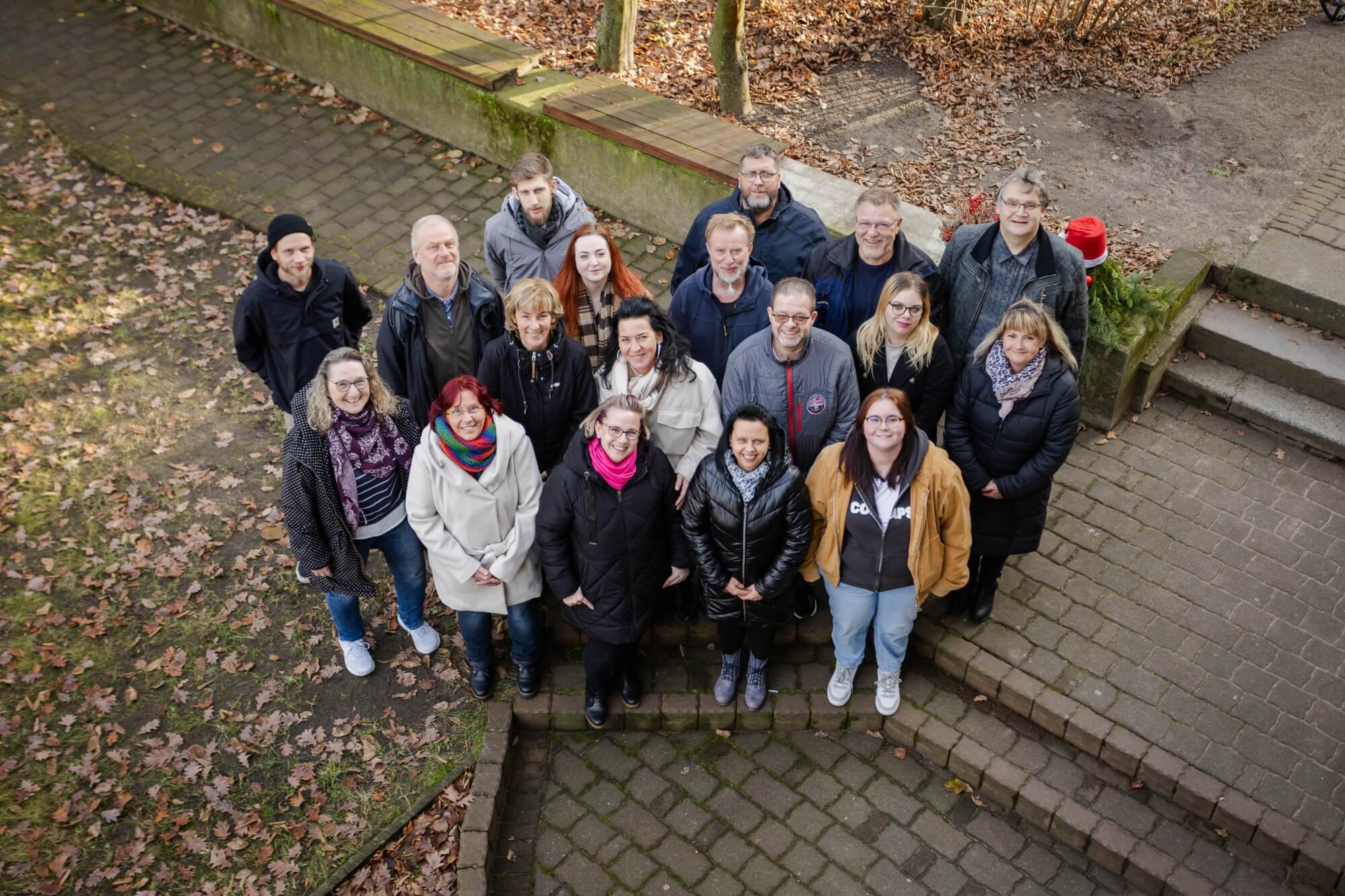 Auf dem Bild sind 18 Personen zu sehen. Fotografiert aus einer erhöhten Position. Sie gucken alle nach oben in die Kamera und stehen auf einem gepflasterten Weg, der von Wiesen gesäumt ist auf denen Blätter liegen.